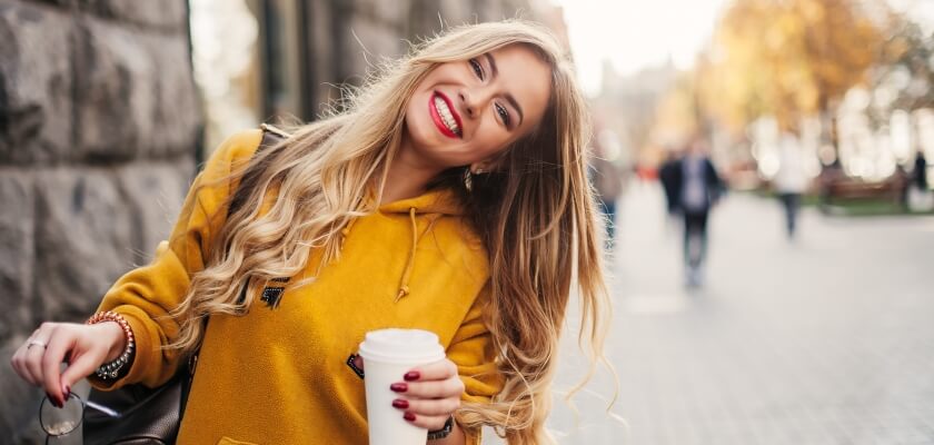 Sieviete priecīga iet pa ielu un rokās tur krūzīti ar kafiju