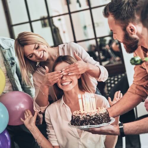 Друзья поздравляют женщину с днем рождения сюрпризом - тортом и воздушными шарами.